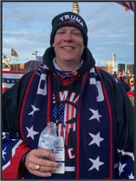 A cleanshaven white man smiling, displaying triple smile lines around his face, wearing a US flag punisher sweatshirt, a scarf or robe with ostentatious US flag stars, and a trump hat or headband.  He is sporting a flat gold ring on his large left hand.