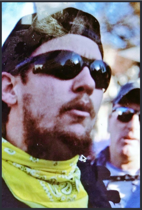 A white man with a brown beard in a weird goatee style wearing bulbous black sunglasses and a backwards ball cap.