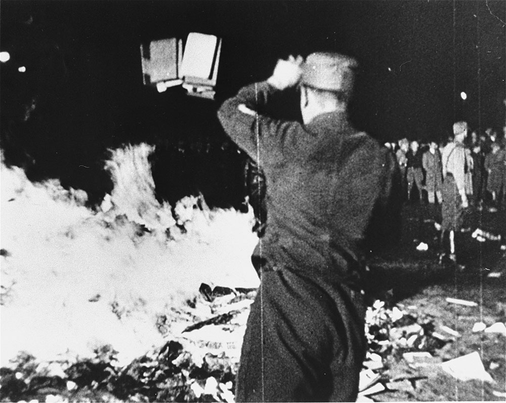 Nazi goon throwing books on a large fire during the 1933 book burnings