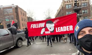 A large bright red banner with the text "Follow Your Leader" and a stylized picture of Hitler blowing out his brains is carried in a crowd of people dressed mostly in black at a Saturday counter-protest.