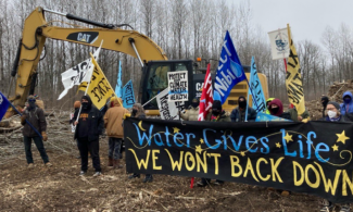 Pipeline resistance organizers holding "Water Gives Life. We Won't Back Down" banner in front of backhoe