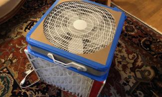 A box fan taped to a box made of filters, with cardboard covering the parts of the fan face where the circle of fan blades do not reach.