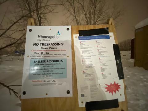 Laminated flyer posted on plywood, with snow and trees in the bright winter night behind it, reading: "Minneapolis, City of Lakes No Trespassing! Please Vacate All tents, structures and personal belongings must be moved Dec-28-22 - Please see below for information about how to access shelters. - Outreach workers are available to provide information and help make service connections. Shelter Resources Adult Shelter Connect (Single Adults) 612-248-2350 Hennepin County Shelter Team (Family Shelter) 612-348